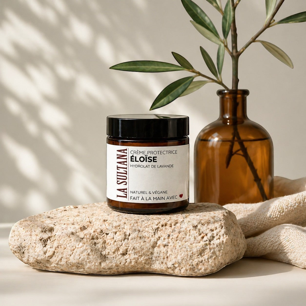 Pot de crème réparatrice ÉLOÏSE blanche à étiquette noire et blanche, posé sur pierre naturelle beige, flacon ambré avec.
