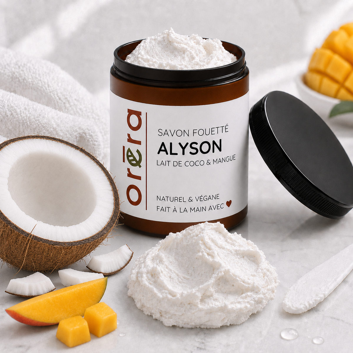 Pot marron foncé de savon fouetté blanc coco et mangue avec étiquette beige, couvercle noir posé à côté.