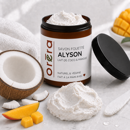 Pot marron foncé de savon fouetté blanc coco et mangue avec étiquette beige, couvercle noir posé à côté.