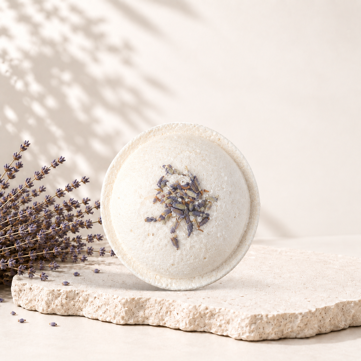 Bombe de bain ronde blanche avec fleurs de lavande séchées sur le dessus, posée sur pierre beige texturée.