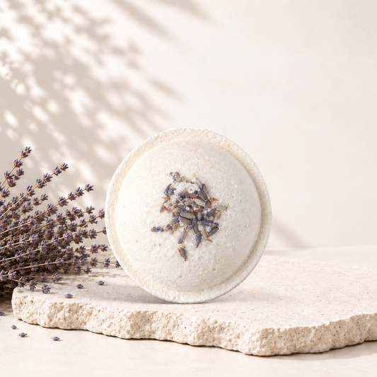Bombe de bain ronde blanche avec fleurs de lavande séchées sur le dessus, posée sur pierre beige texturée.