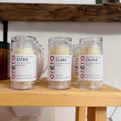 Trois baumes à lèvres Oréra en tube transparent avec étiquettes crème et rouge, présentés sur une étagère en bois.
