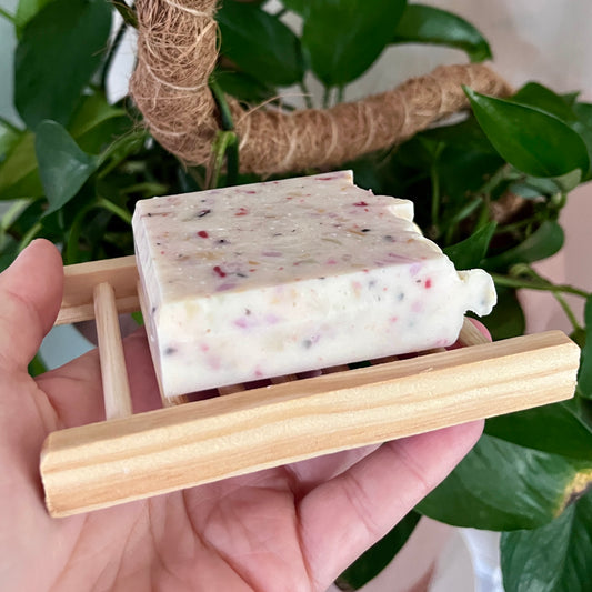 Porte savon en bois naturel tenant un savon blanc moucheté de pétales séchés, compact et artisanal.