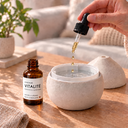 Flacon brun avec étiquette blanche Synergie Vitalité à côté d’un diffuseur blanc, goutte d’huile essentielle tombant d’un.