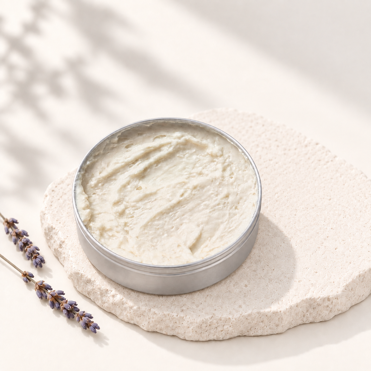 Baume féminin crème blanche dans pot rond en aluminium sur support texturé beige avec brins de lavande sèches.