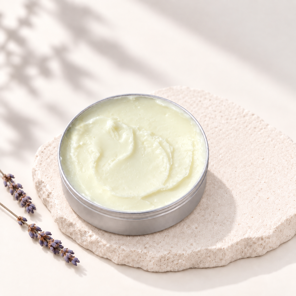 Baume à tatouage jaune pâle dans pot rond en métal posé sur pierre beige texturée avec brin de lavande sèche.
