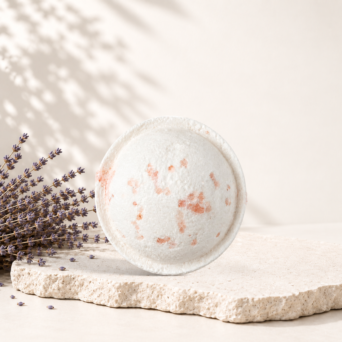 Bombe de bain ronde blanche avec éclats rouges, style artisanal, posée sur pierre beige avec lavande sèche.