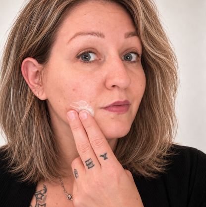 Femme appliquant crème hydratante blanche sur joue, peau claire, cheveux blonds.