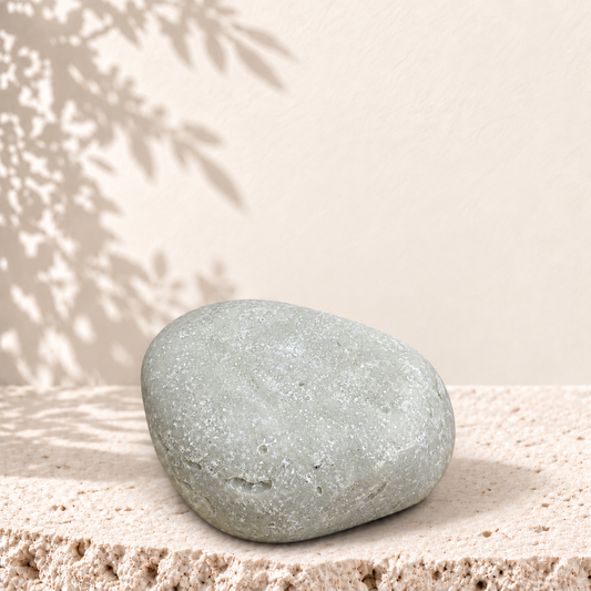 Savon exfoliant gris clair ovale avec texture pierre ponce posée sur surface beige texturée.