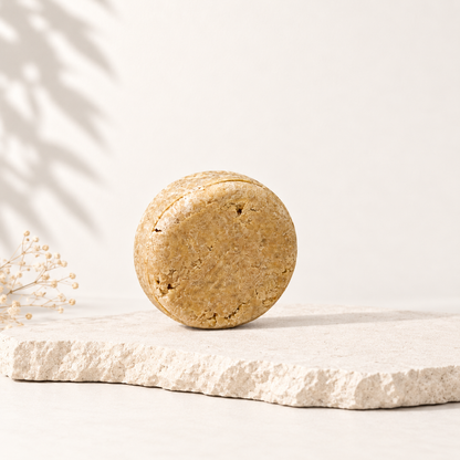 Shampoing solide rond beige en texture naturelle posé sur pierre brute, ambiance épurée.