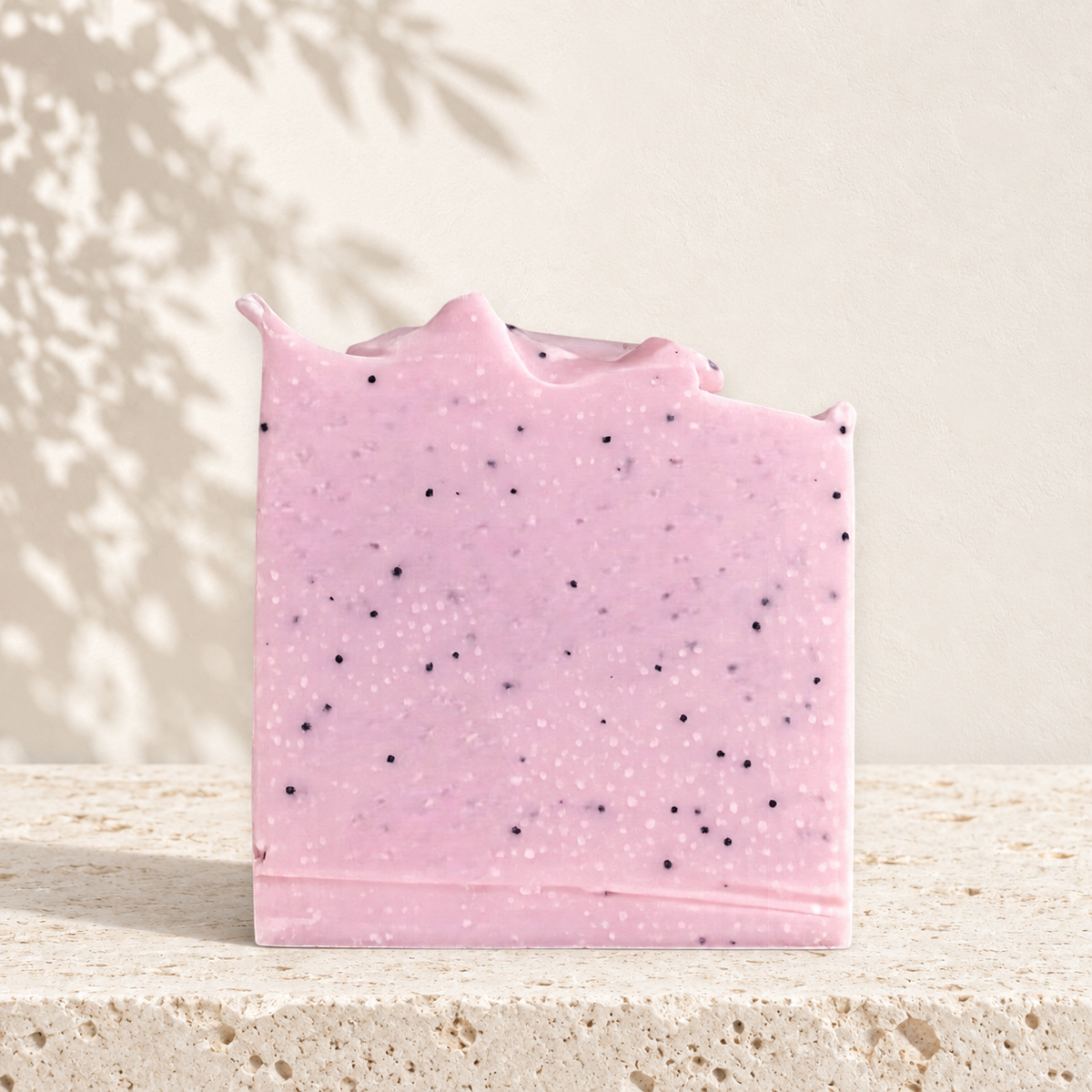Savon exfoliant rose pâle avec grains noirs, forme carrée irrégulière, texture riche.