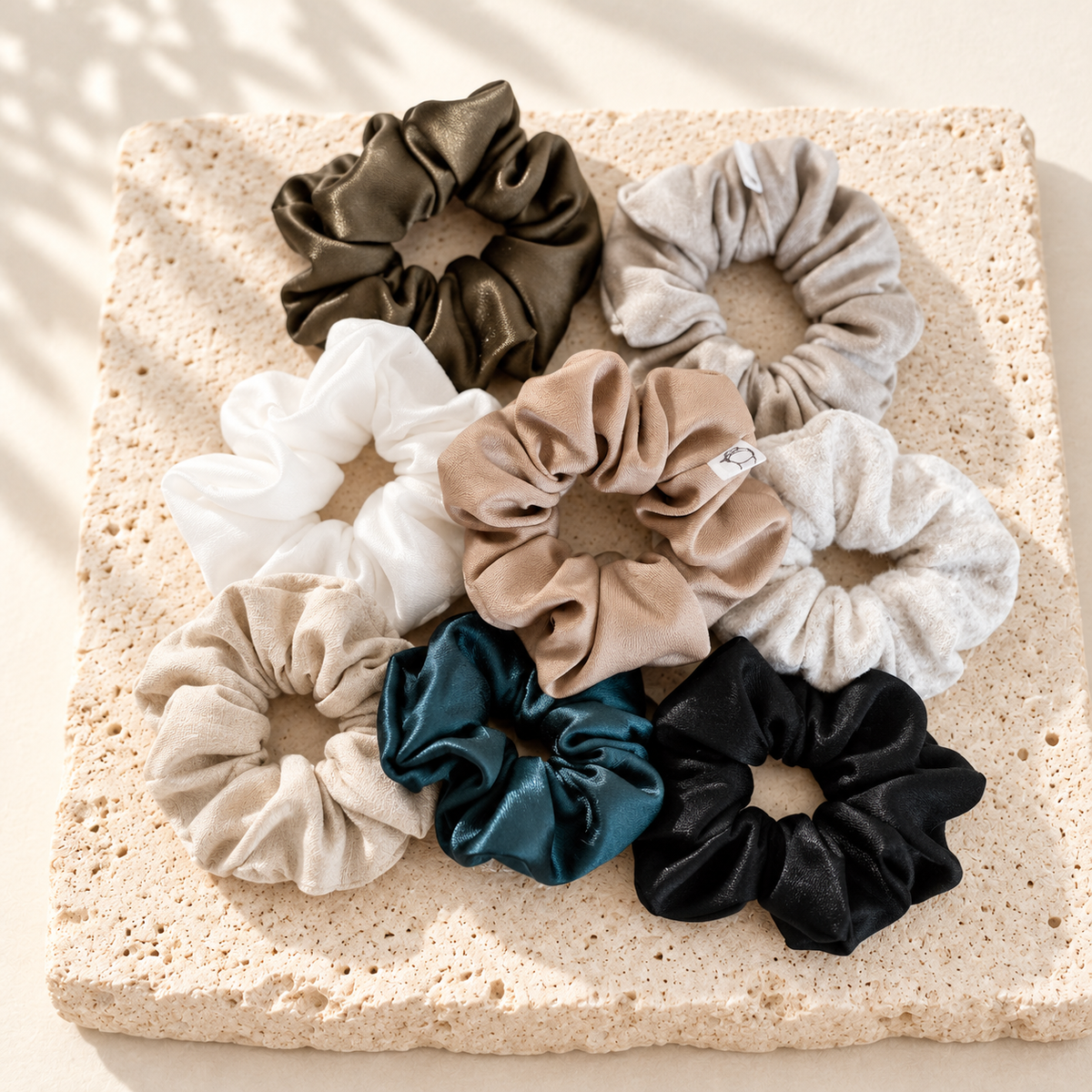 Assortiment de scrunchies en tissus et couleurs variés, posés sur du travertin clair.