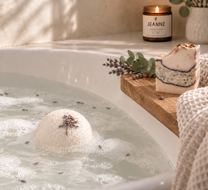 Bombe de bain blanche dans l’eau mousseuse, avec une bougie JEANNE et un savon sur un plateau en bois.