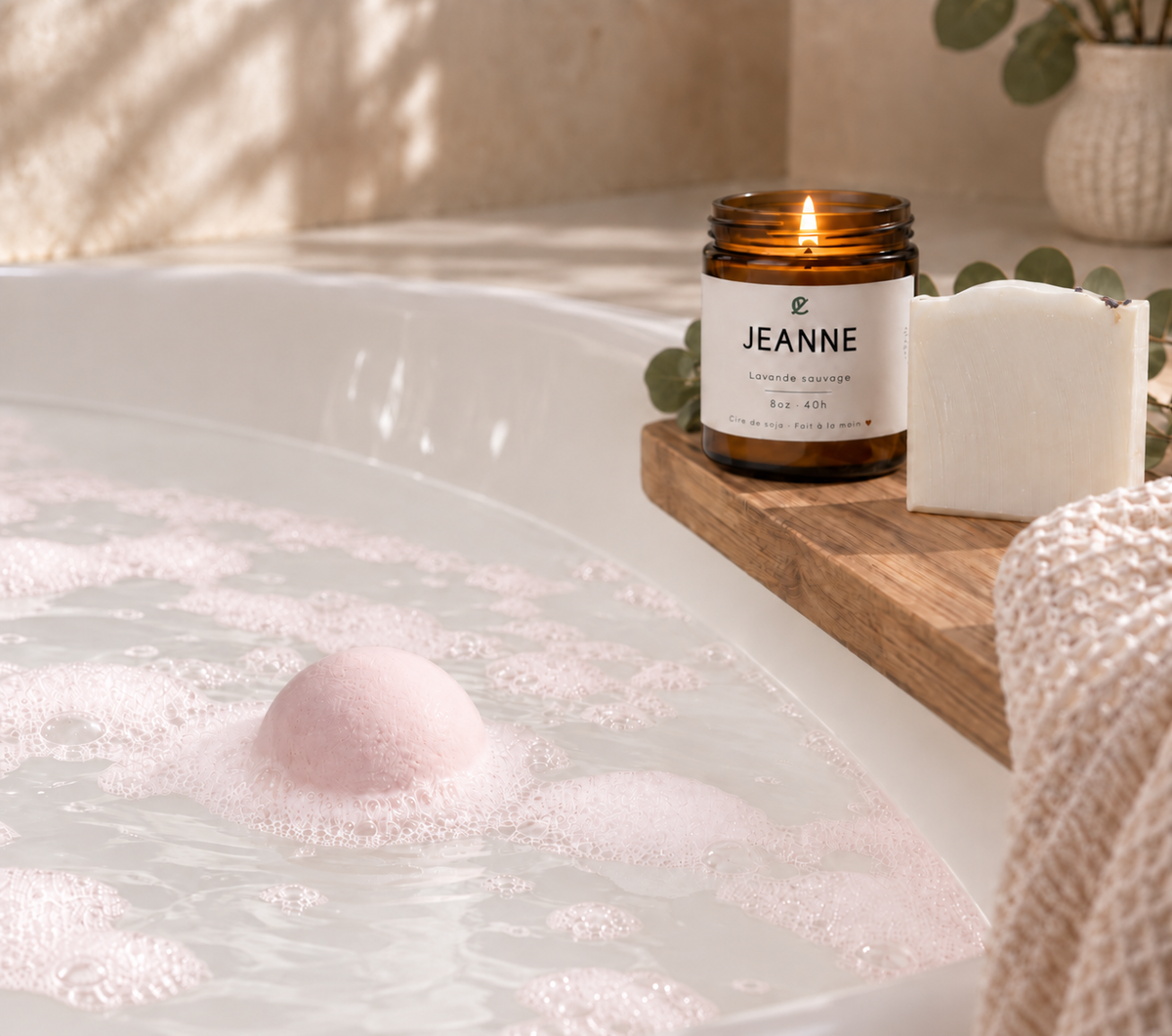 Bombe de bain ronde rose pâle effervescente dans baignoire blanche avec bougie et savon sur plateau en bois.
