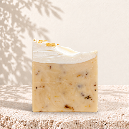 Savon NÉFERTITI Oréra beige aux pétales de calendula, surmonté d’une couche blanche ondulée, sur une pierre claire.