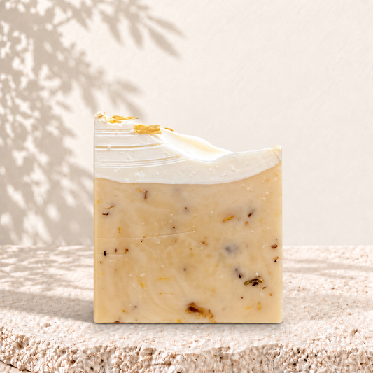 Savon NÉFERTITI Oréra beige aux pétales de calendula, surmonté d’une couche blanche ondulée, sur une pierre claire.