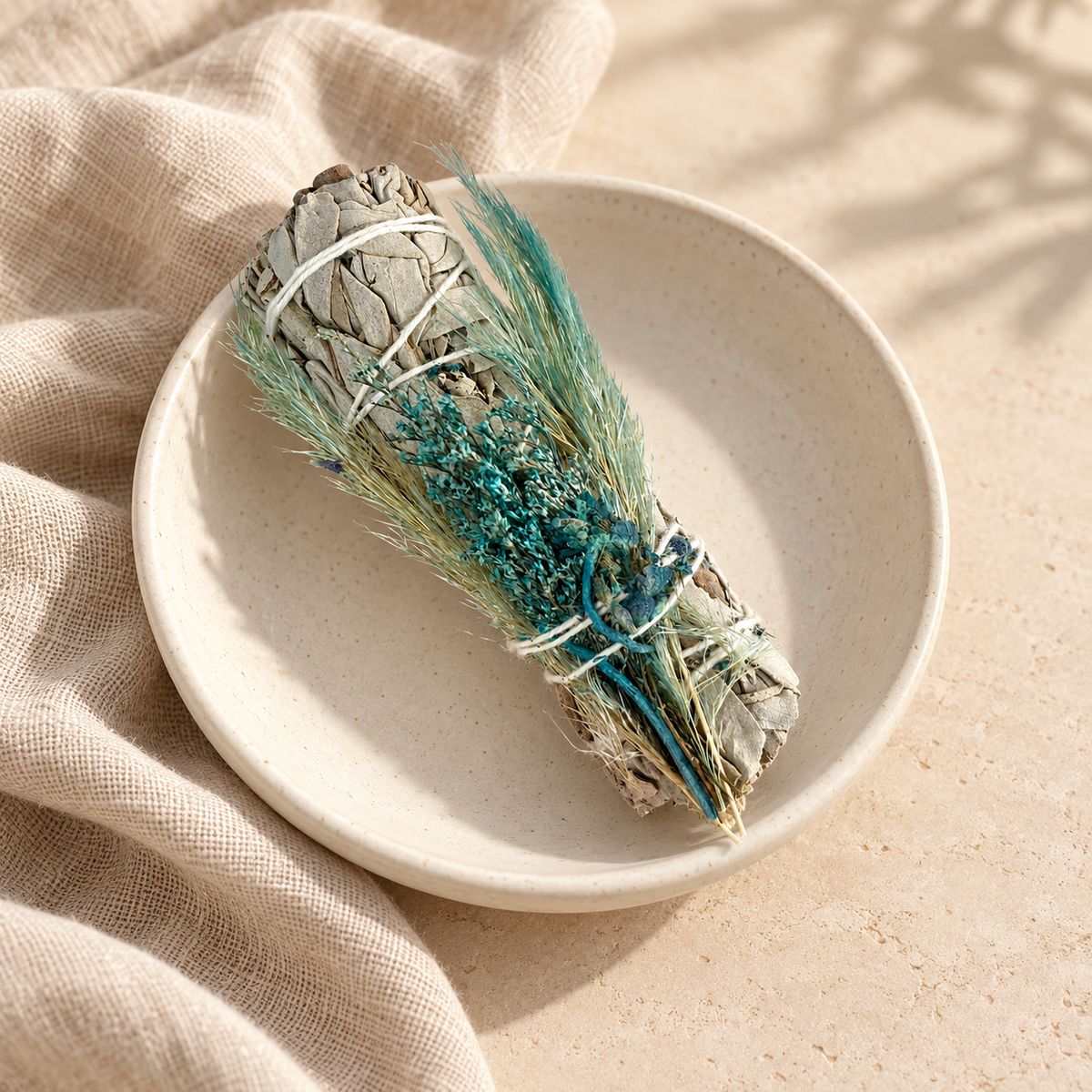 Bâton de sauge blanche florale bleu-vert, non allumé, dans une coupelle en céramique ivoire avec du lin beige.