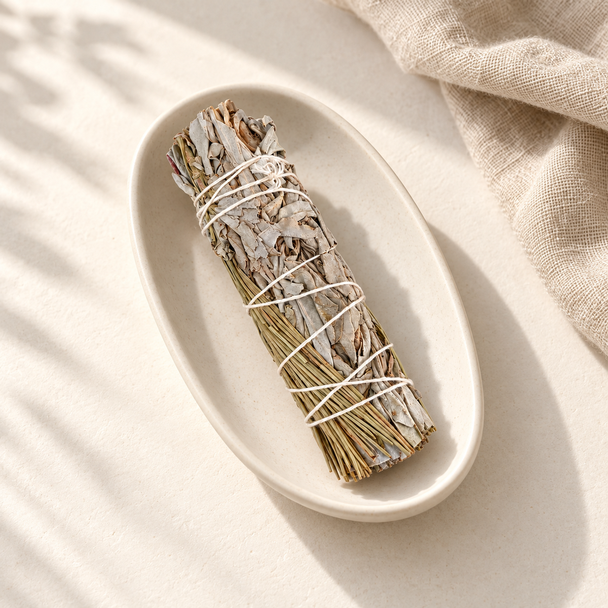 Bâton de sauge blanche et pin, non allumé, dans une coupelle en céramique ivoire avec du lin beige.