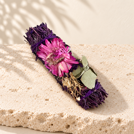 Bâton de sauge lavande et fleurs séchées rose-violet, couché sur une dalle de pierre claire.