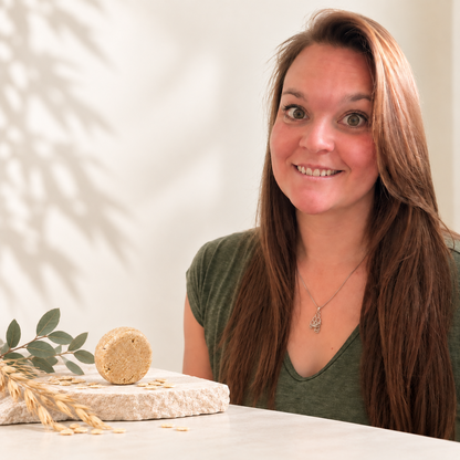 Femme souriante aux cheveux longs, à côté d’un shampoing solide rond beige posé sur une pierre.