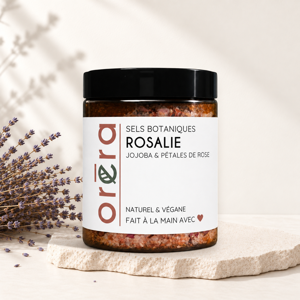 Pot en verre avec sels botaniques rosés, pétales de rose visibles, étiquette minimaliste blanche, couvercle noir.