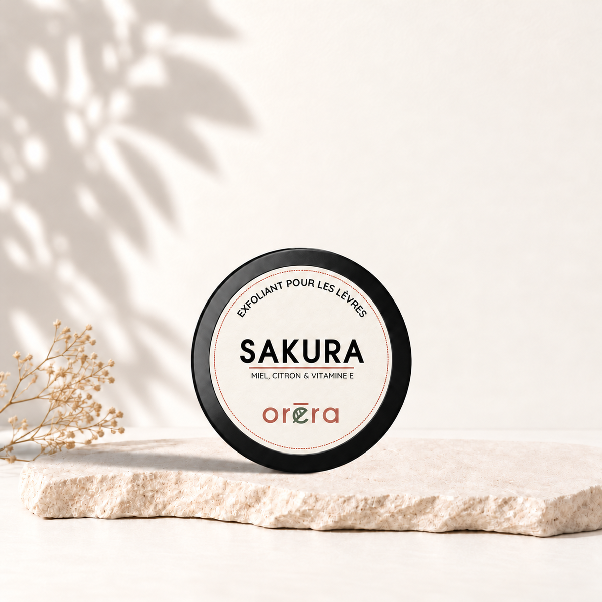 Pot rond noir SAKURA avec nouvelle étiquette ORÉRA, exfoliant pour les lèvres au miel, citron et vitamine E, sur une pierre beige.