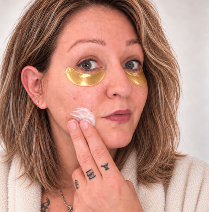 Femme appliquant crème de nuit blanche, patchs oculaires dorés sous les yeux, peau claire, cheveux châtains.