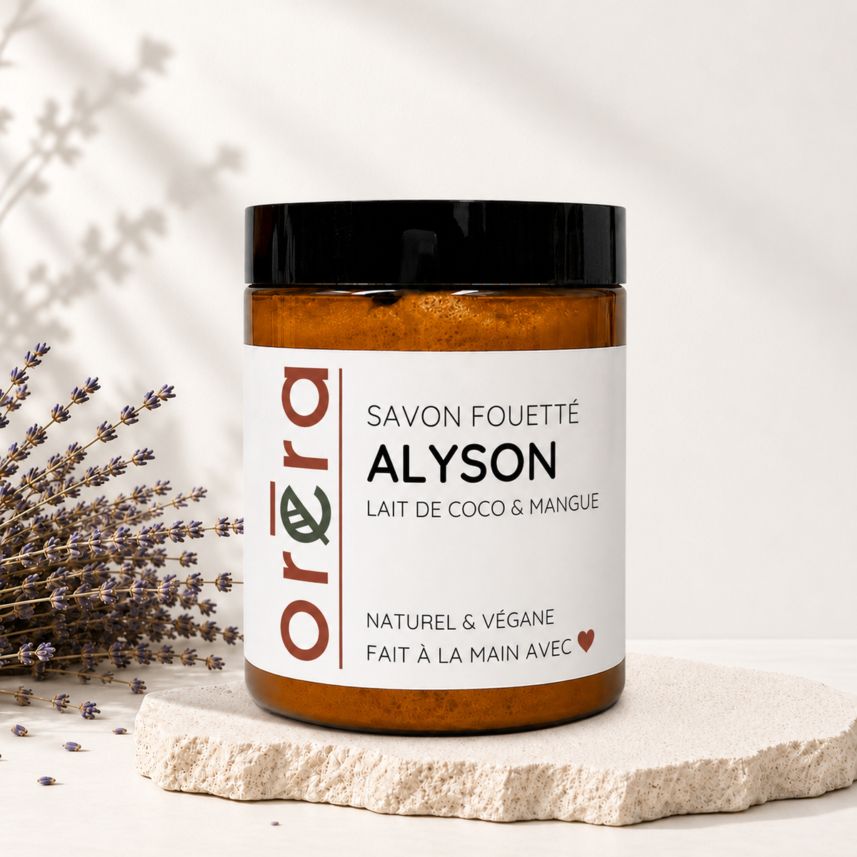 Pot en verre ambré avec couvercle noir, étiquette blanche indiquant savon fouetté Alyson coco et mangue, style minimaliste.