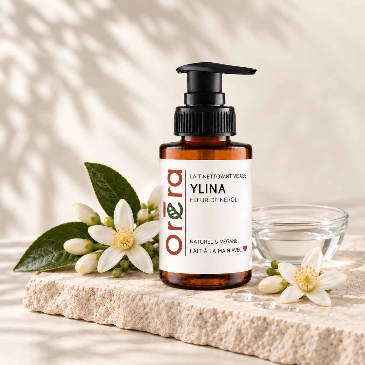 Flacon-pompe ambré de lait nettoyant Ylina à la fleur de néroli, entouré de fleurs blanches et d’un bol transparent.